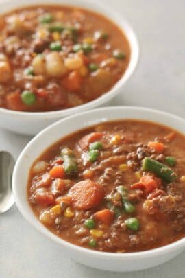 Two bowls of hearty beef and vegetable soup with visible pieces of carrots, potatoes, green beans, peas, and corn. A silver spoon is placed beside the bowls on a light surface.