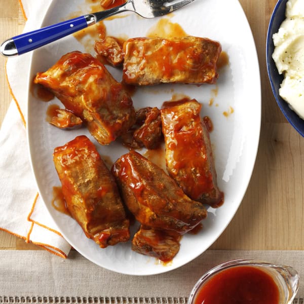 Plate of barbecue-glazed ribs with a fork, served next to a bowl of mashed potatoes and a pitcher of sauce on a wooden surface.