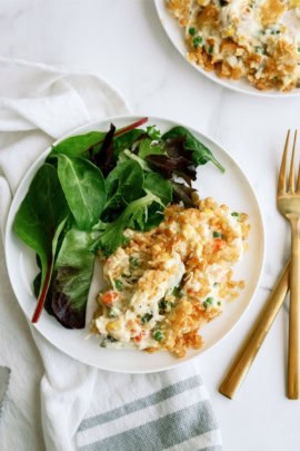 A white plate with a serving of vegetable casserole topped with crispy bits, accompanied by mixed green salad leaves. A cloth napkin and gold-colored fork and knife are placed beside the plate.