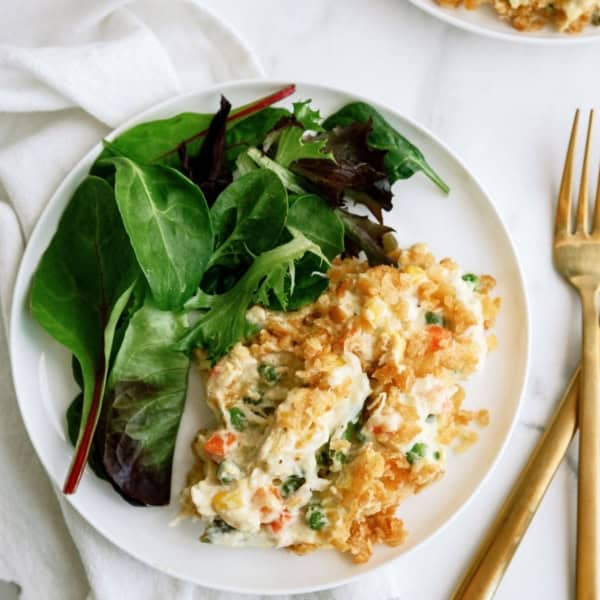 A white plate with a serving of vegetable casserole topped with crispy bits, accompanied by mixed green salad leaves. A cloth napkin and gold-colored fork and knife are placed beside the plate.