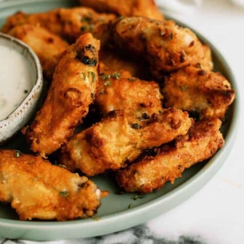 A plate of crispy fried chicken wings with a bowl of dipping sauce on the side, placed on a checkered cloth.