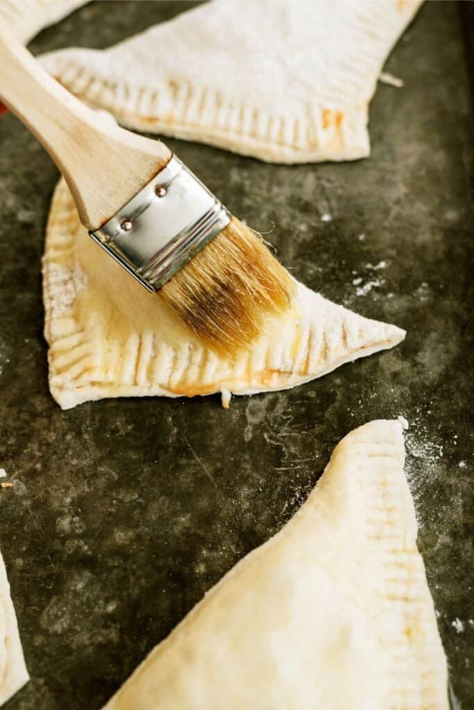 A pastry brush brushing egg white on top of an uncooked Calzone on a baking sheet.