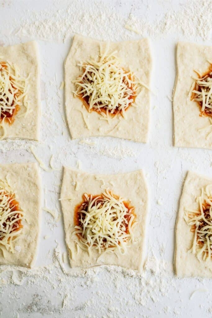 6 pizza dough squares with sauce and cheese centered in each square.