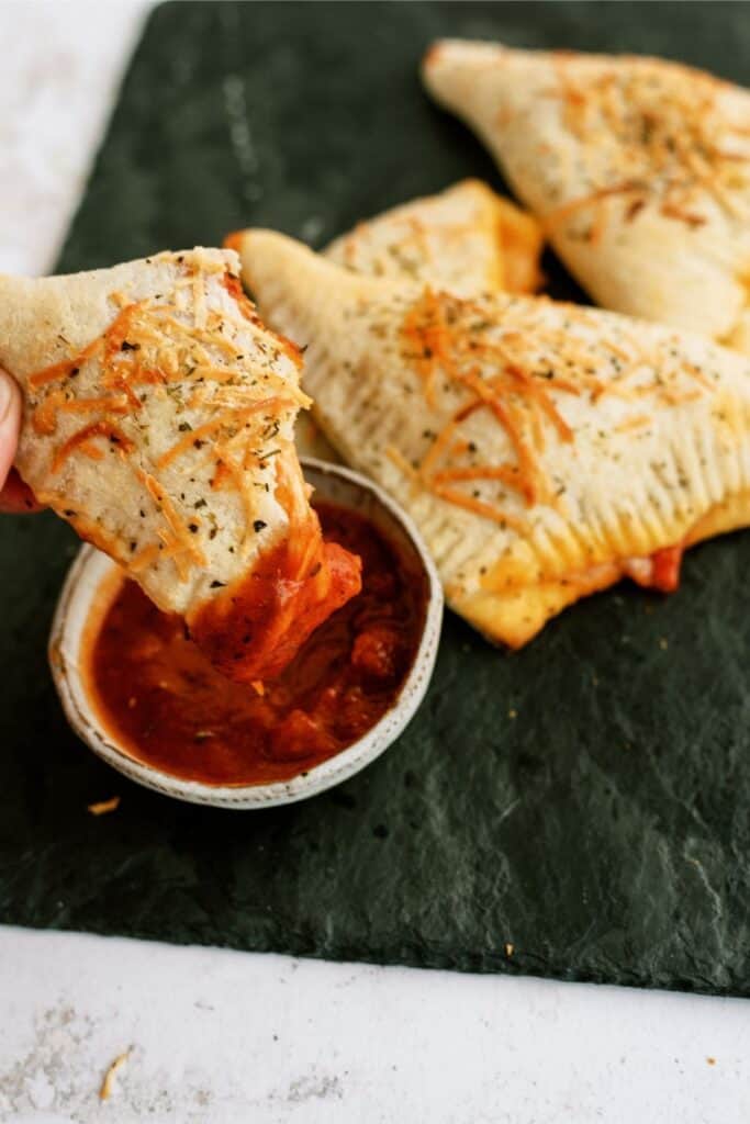 A hand dipping one Quick and Easy Calzones in a cup of sauce with other Quick and Easy Calzones in the background.