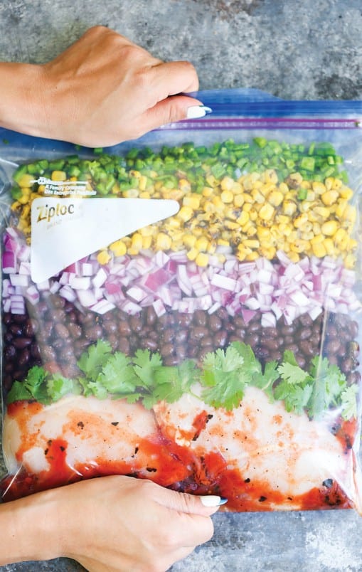 Hands holding a partially sealed plastic bag containing raw chicken, cilantro, black beans, red onions, corn, and green bell peppers, arranged in layers.