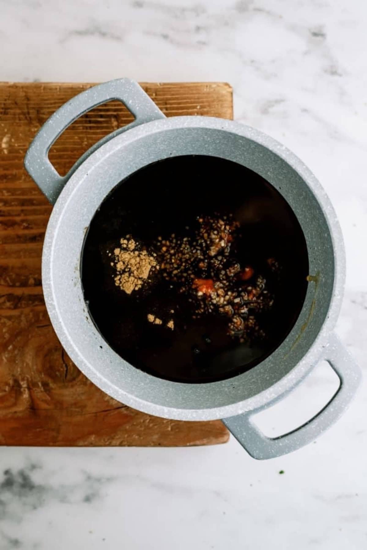 Pot filled with dark liquid and spices, placed on a wooden board on a marble surface.