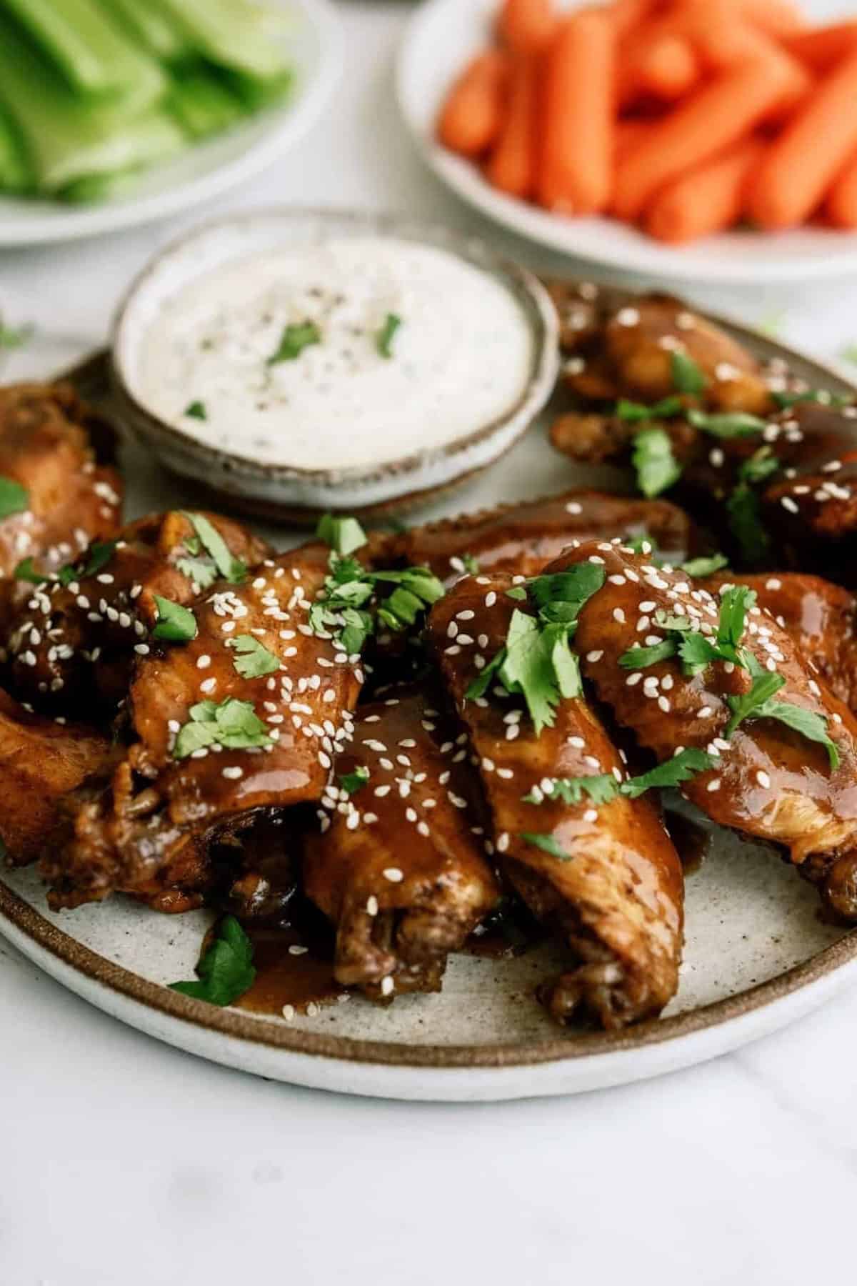 Plate of glazed chicken wings garnished with sesame seeds and herbs, accompanied by a bowl of creamy dip. Plates of celery and carrot sticks in the background.