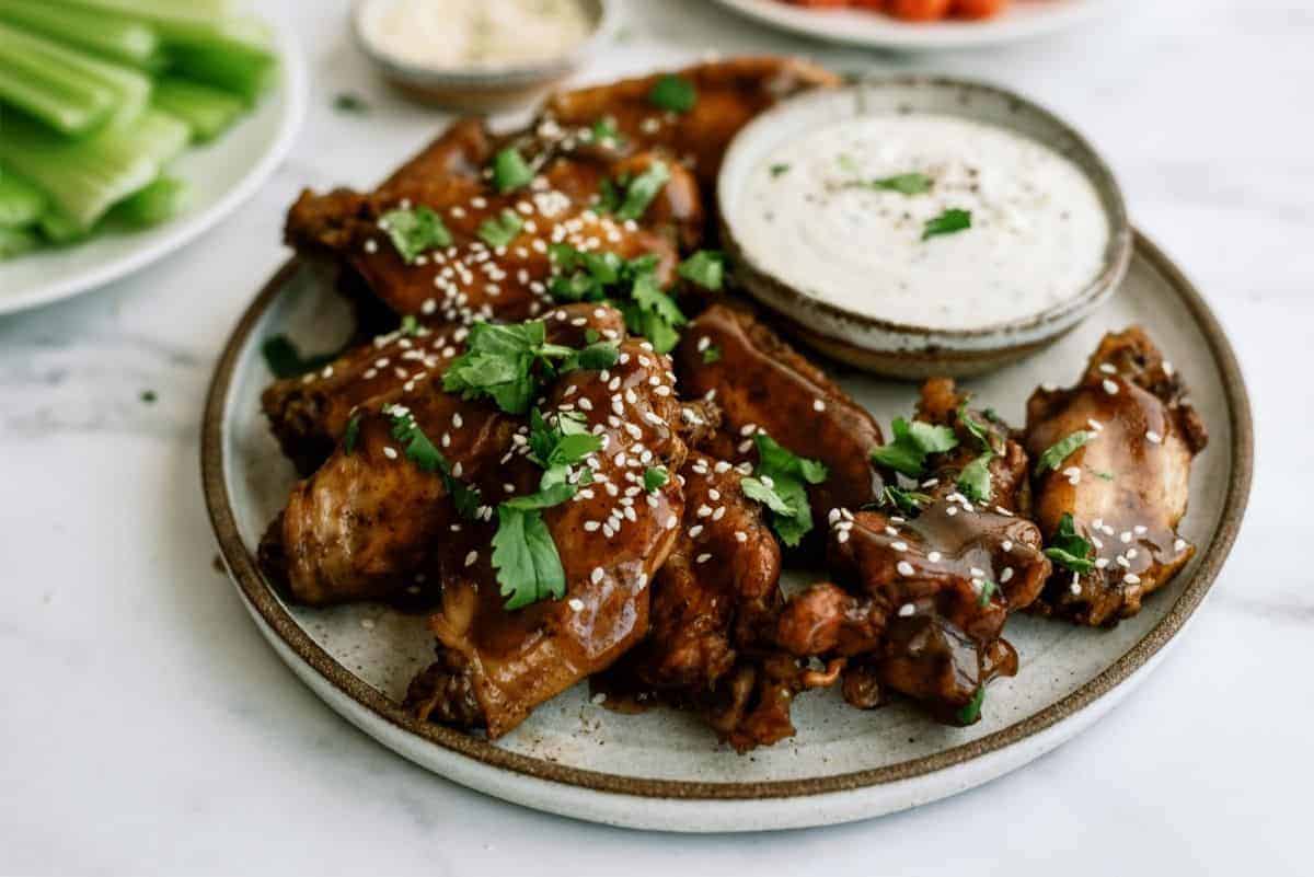 A plate of saucy chicken wings garnished with sesame seeds and cilantro, served with a side of dip.