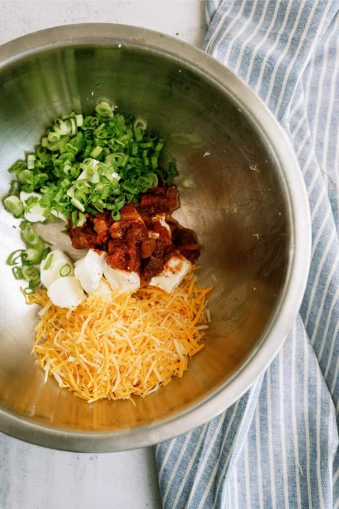 A metal bowl contains chopped green onions, chunks of meat, sour cream, cream cheese, and shredded cheddar, next to a blue striped cloth.