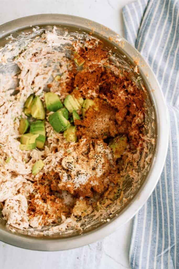 Metal bowl with shredded chicken, diced avocado, spices, and seasoning, next to a blue-striped cloth.