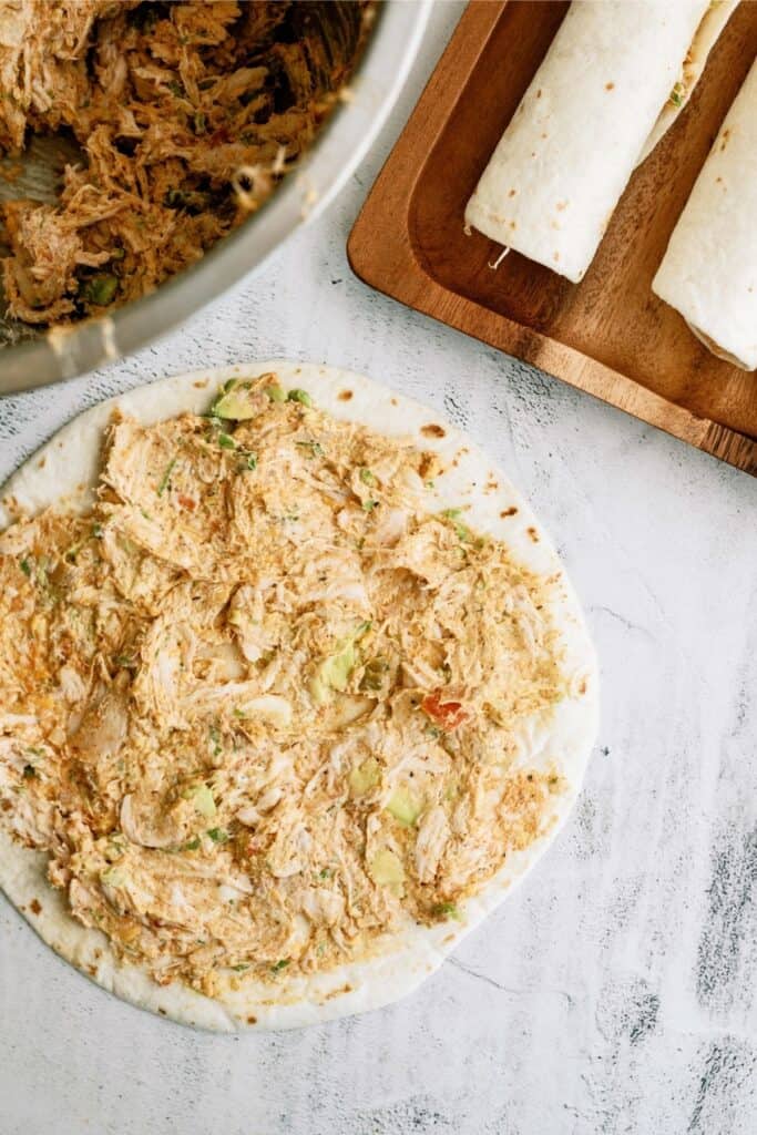 Tortilla spread with shredded chicken mix next to rolled tortillas on a wooden tray. Bowl of chicken mix partially visible.