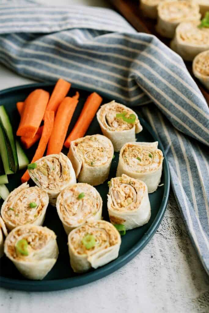 A plate with pinwheel wraps and carrot and cucumber sticks, next to a blue-striped cloth.