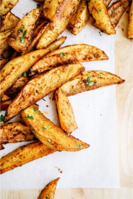 Seasoned potato wedges garnished with parsley on a sheet of parchment paper on a wooden surface.