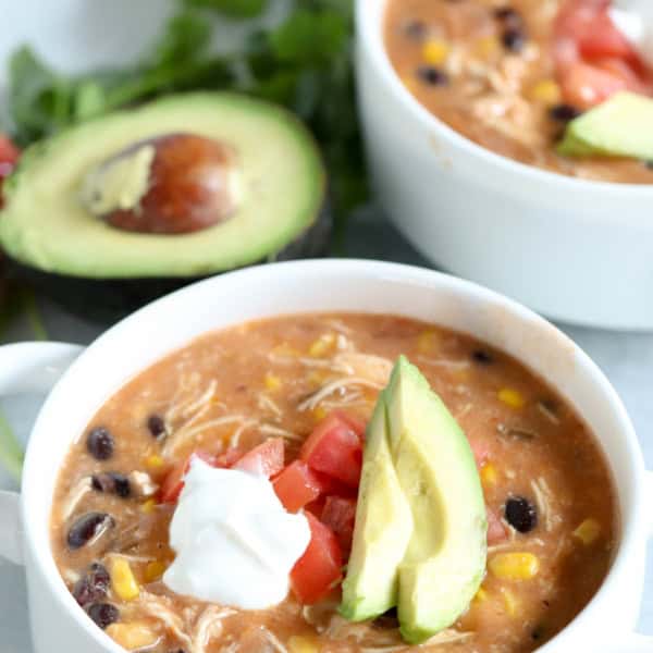 A close-up of two bowls of soup, garnished with avocado slices, diced tomatoes, and a dollop of sour cream. An avocado cut in half and some greens are in the background.