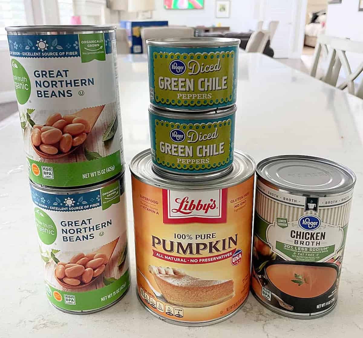 Cans of Great Northern Beans, diced green chile peppers, pure pumpkin, and chicken broth are arranged on a kitchen countertop.