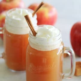 Two glass mugs of apple cider topped with whipped cream and garnished with cinnamon sticks, with apples in the background.