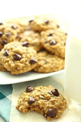 A plate of chocolate chip cookies with a glass of milk on the side. One cookie in the foreground has a bite taken out of it.