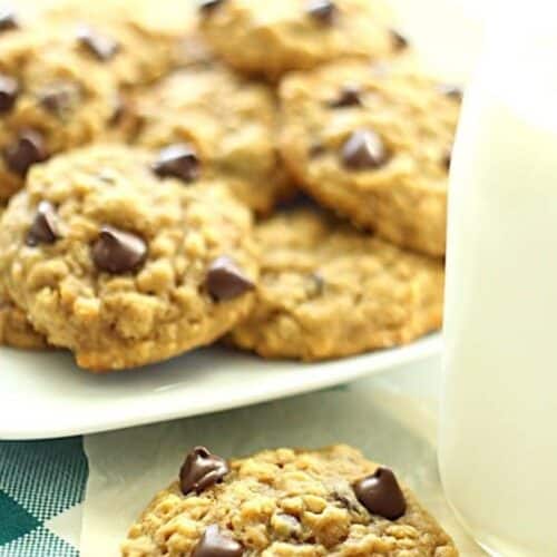 A plate of chocolate chip cookies with a glass of milk on the side. One cookie in the foreground has a bite taken out of it.