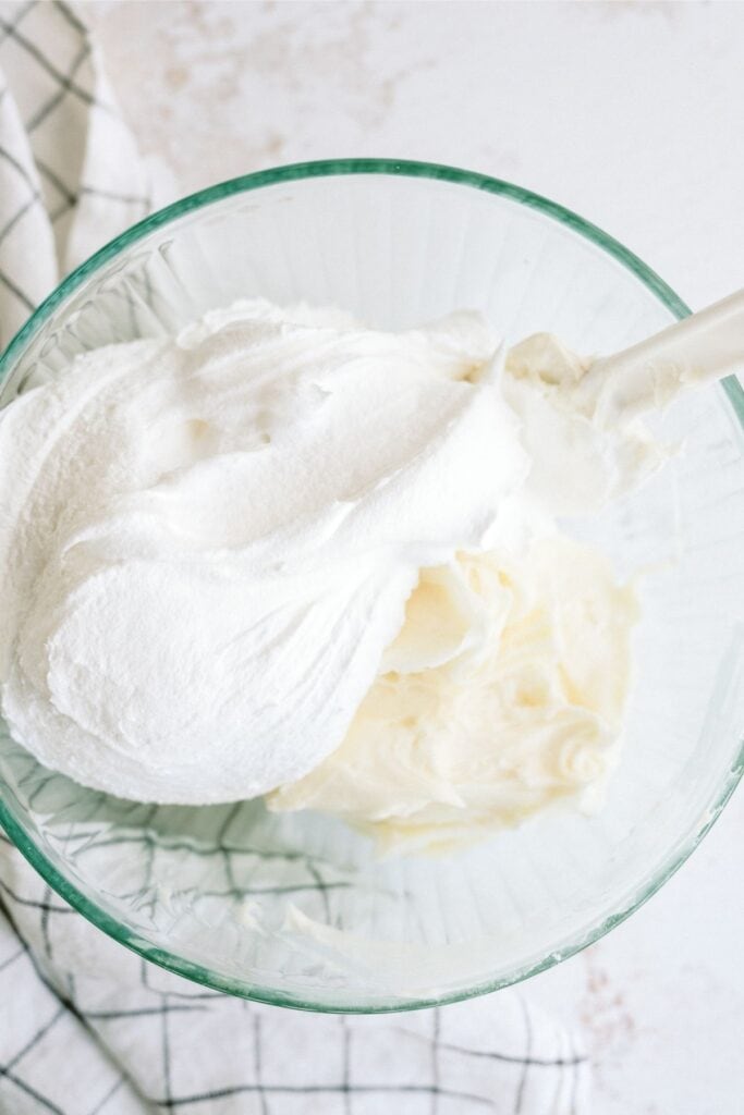 A bowl containing fluffy white whipped cream being mixed with a spatula, placed on a cloth with a checkered pattern.