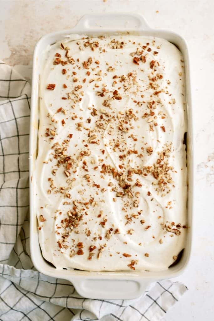 A rectangular baking dish filled with frosted cake topped with sprinkled chopped nuts. A white and green checkered cloth is partially visible on the left side.