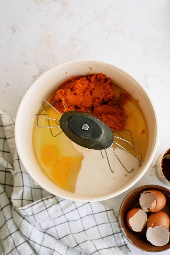 A mixing bowl contains pumpkin puree, eggs, and sugar with beaters resting on top. Next to the bowl are cracked eggshells in a small bowl, placed on a cloth napkin.