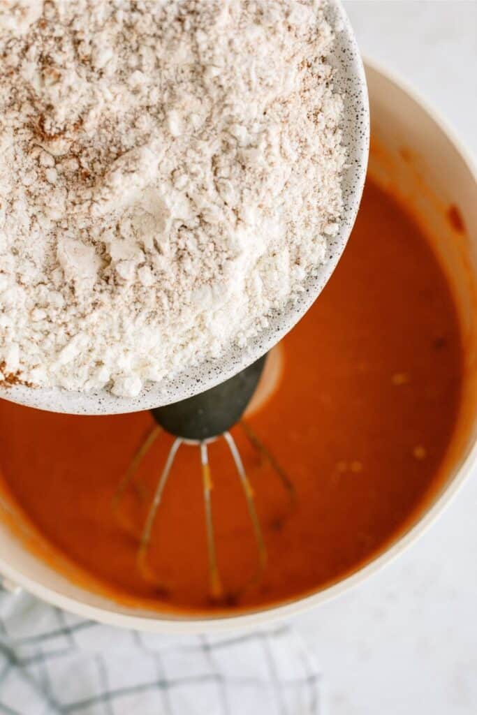 Close-up of flour being poured from a white bowl into a mixing bowl containing a thick orange batter, with a black whisk partially submerged in the batter.