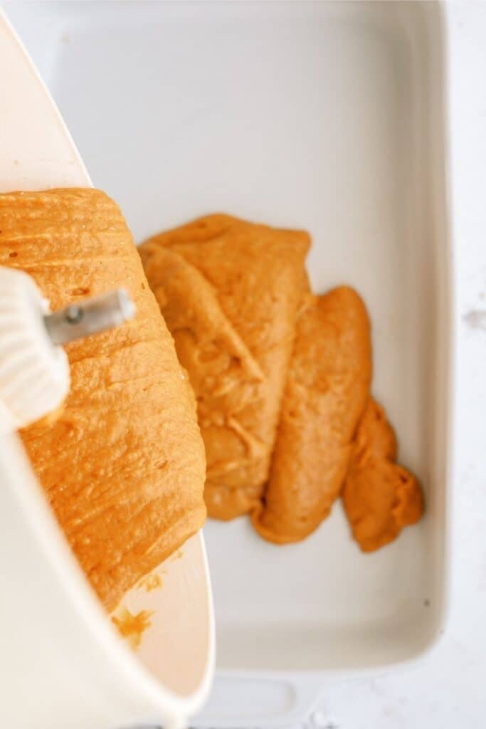 A white mixing bowl is pouring an orange batter into a white rectangular baking dish.