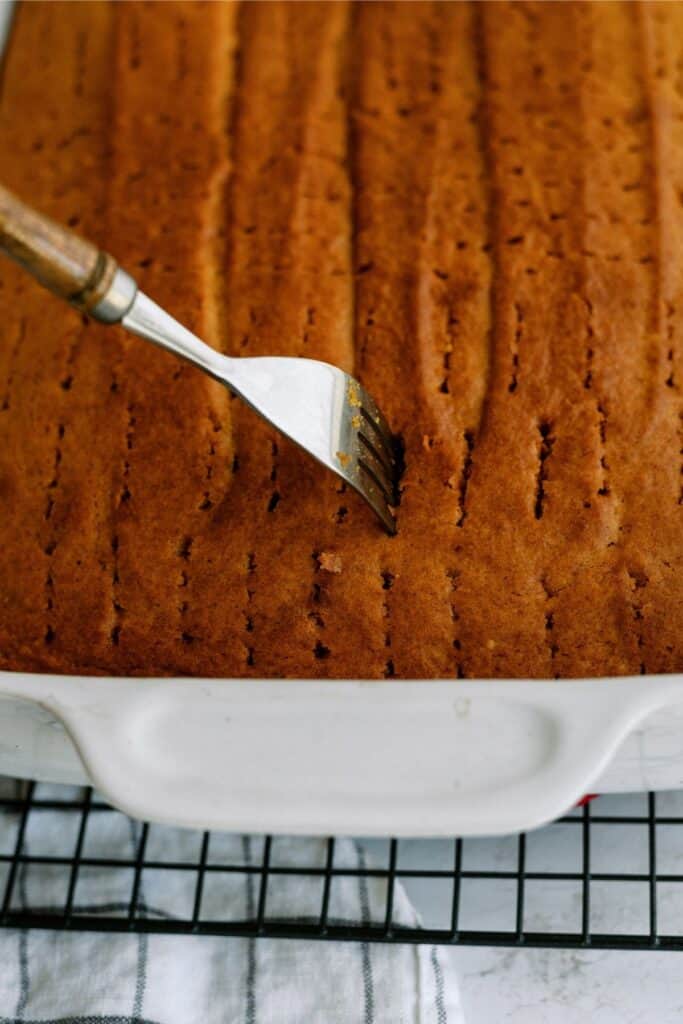 A fork is used to poke holes in a golden-brown cake resting in a white baking dish on a cooling rack.