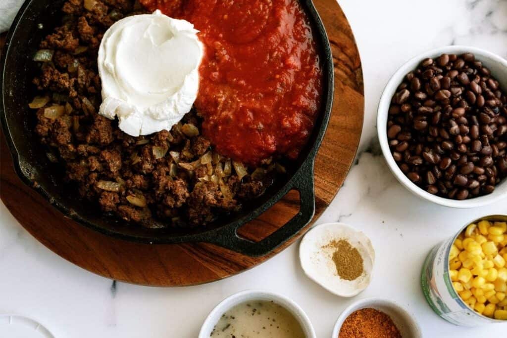 A skillet with ground beef, onions, sour cream, and tomato sauce. Surrounding bowls contain black beans, corn, spices, and seasoning. All items are arranged on a wooden board and white surface.