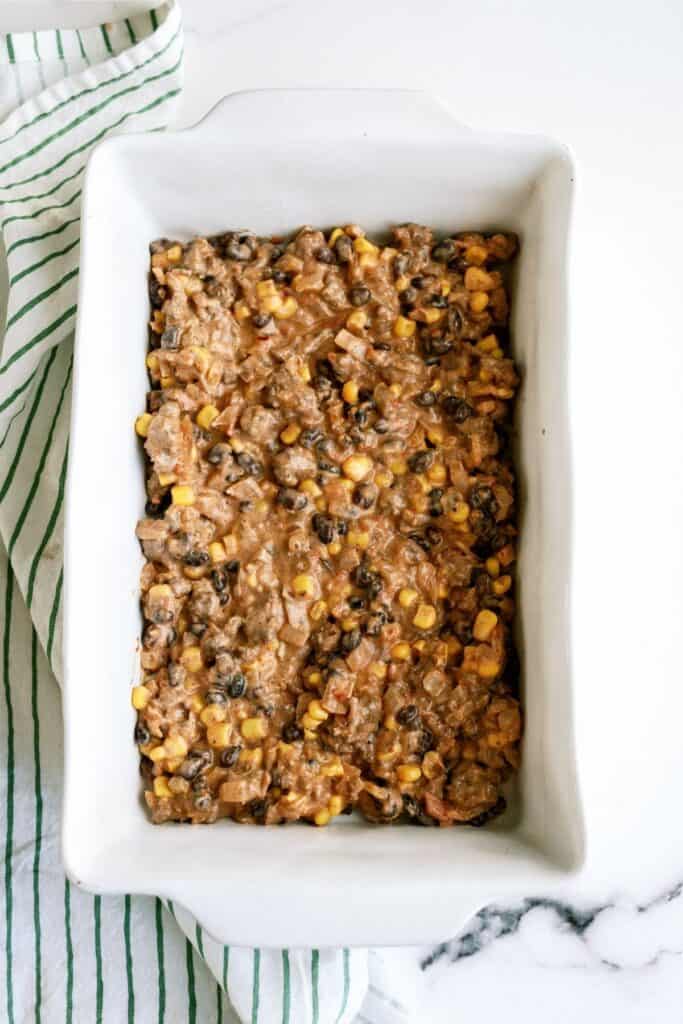 A white rectangular baking dish filled with a mixture of beans, corn, and a creamy sauce is placed on a white surface next to a striped cloth.