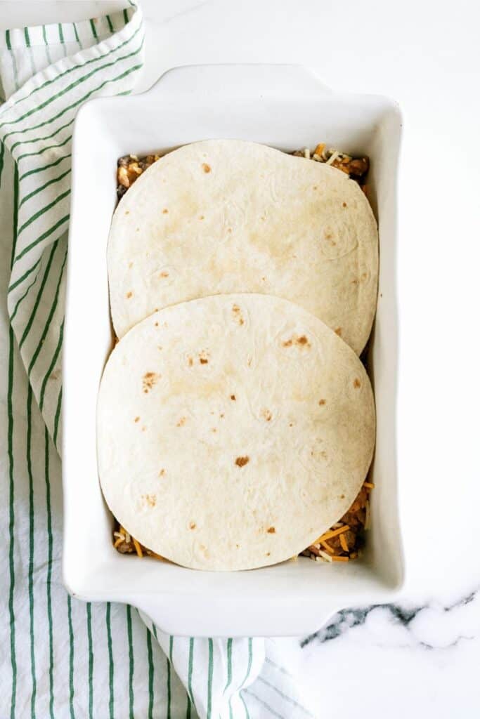 Two flour tortillas placed over a layer of food inside a white rectangular baking dish, which is on a white surface with a green-striped cloth beside it.