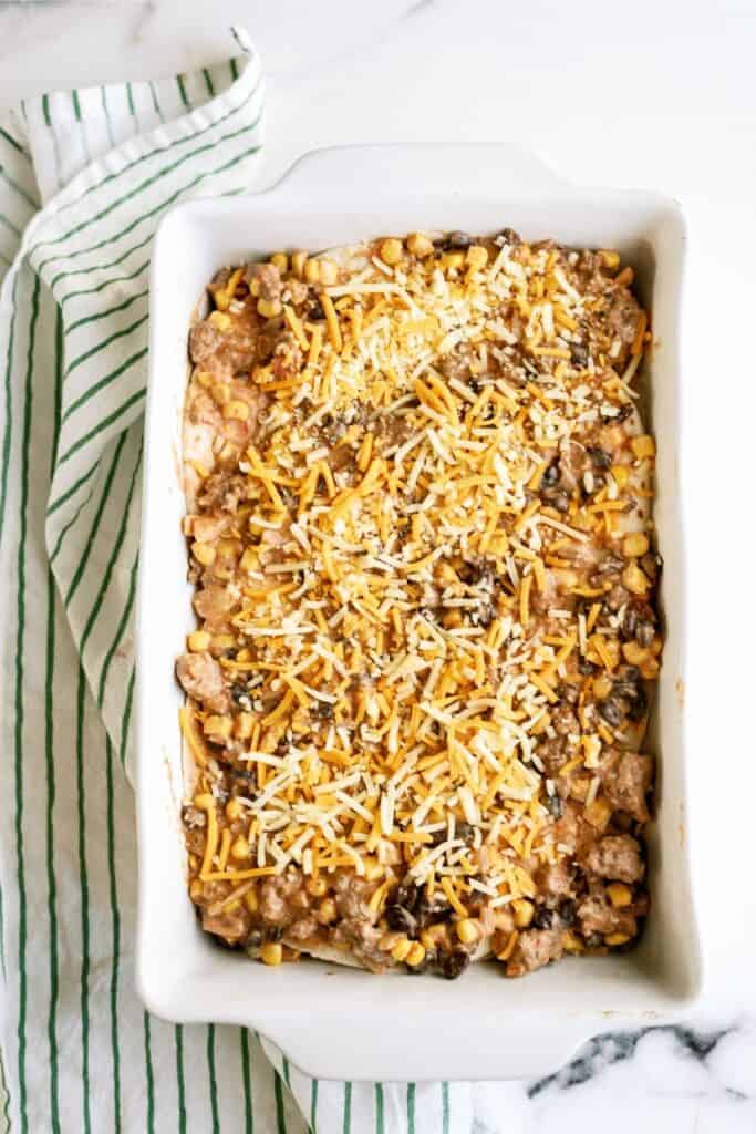 A rectangular baking dish filled with a layered casserole topped with shredded cheese, corn, and black beans, set on a striped cloth.