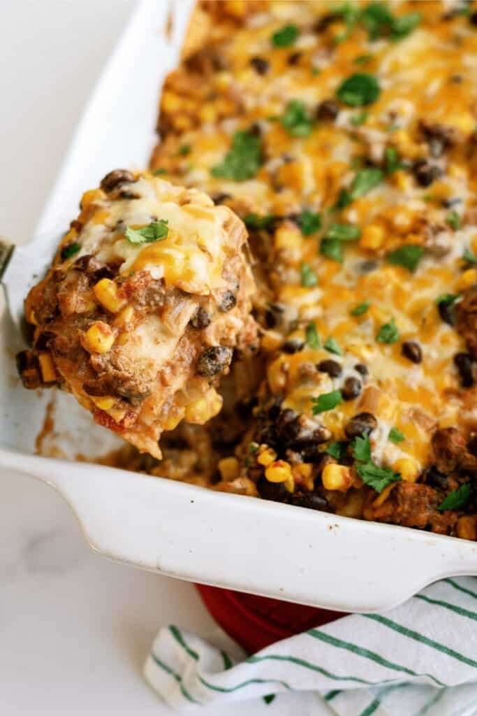 A casserole dish filled with a cheesy, baked mixture of corn, black beans, and meat. A portion is being lifted out with a spatula. The dish is garnished with chopped cilantro.