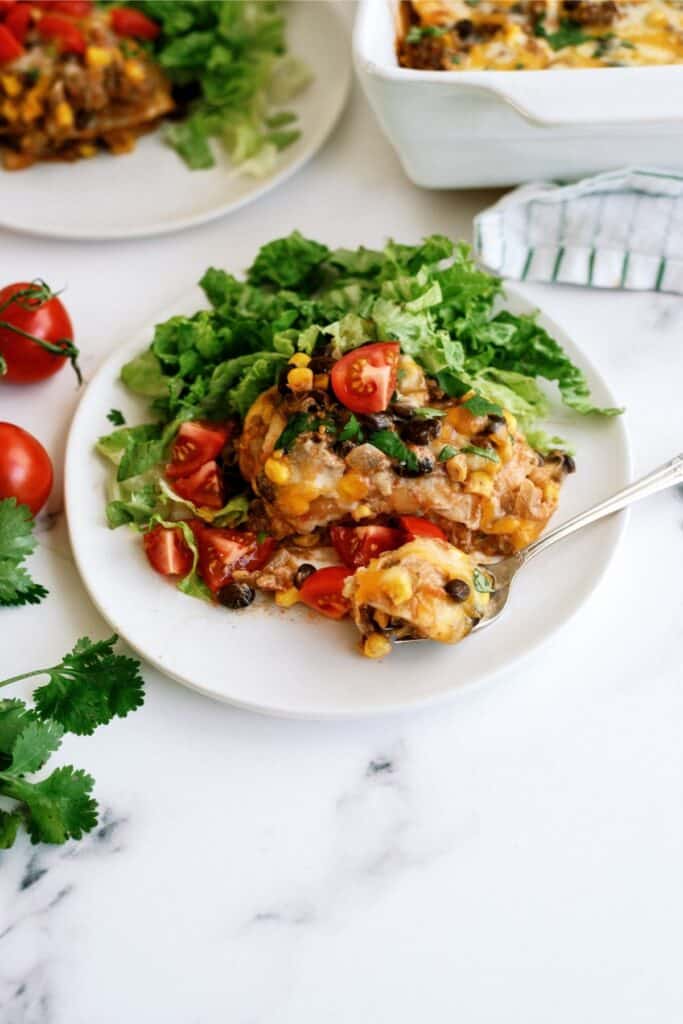 A plate of casserole topped with melted cheese, black beans, corn, chopped tomatoes, and lettuce sits on a white surface, next to fresh tomatoes and cilantro. A utensil rests on the plate.