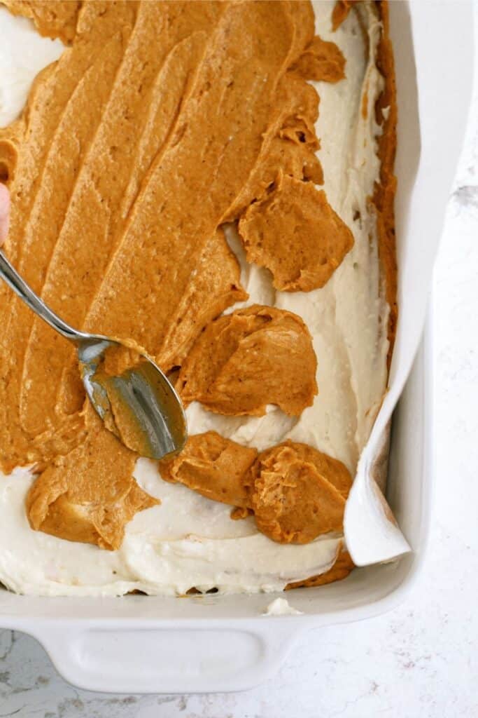 A spoon spreads a layer of brown batter over white batter in a parchment-lined baking dish.