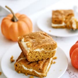 Two pumpkin cream cheese bars are stacked on a white plate with a small pumpkin nearby. Another plate with a bar and a section of a sliced bar is visible in the background.