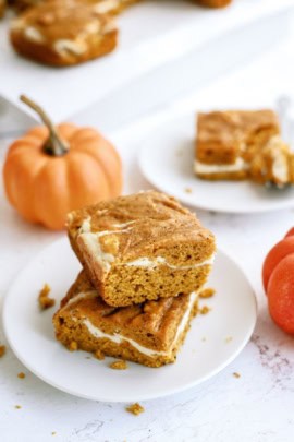 Two pumpkin cream cheese bars are stacked on a white plate with a small pumpkin nearby. Another plate with a bar and a section of a sliced bar is visible in the background.