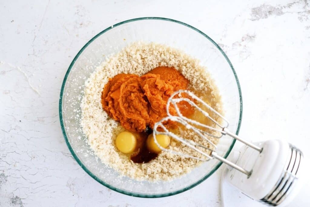 A glass bowl containing ingredients for a recipe, including two eggs, pumpkin puree, and dry mix, is being mixed with an electric hand mixer on a white countertop.