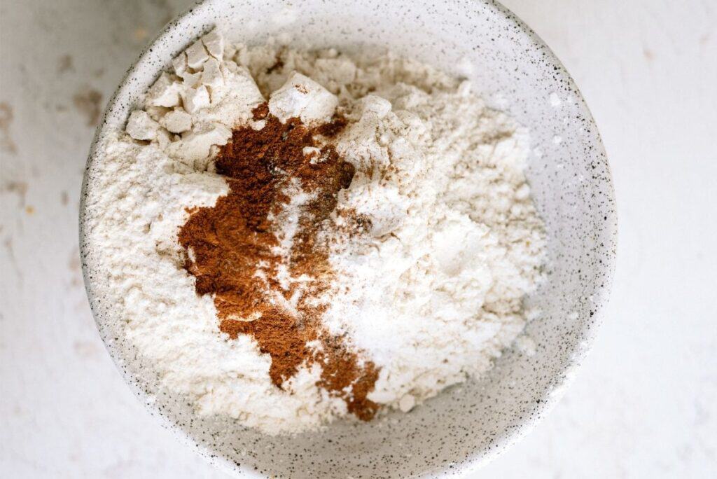 A bowl containing a mix of white flour, brown cinnamon, and white sugar on a white surface.