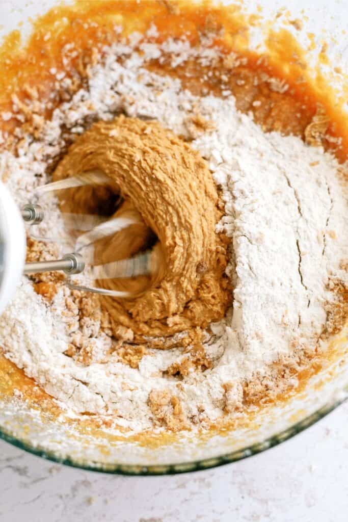 An electric mixer blending flour and a brown mixture in a glass bowl.