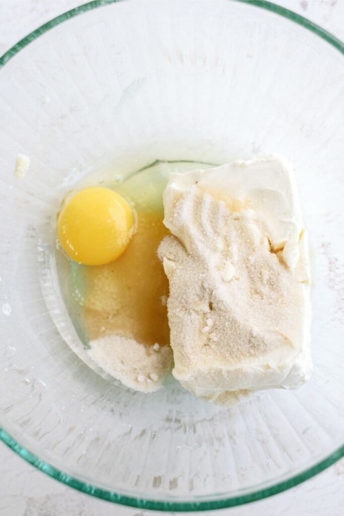 A glass bowl containing an egg, a block of cream cheese, sugar, and vanilla extract.