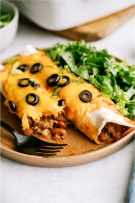 Two Hearty Ground Beef Enchiladas topped with olives on a plate with a salad on the side. One of the enchiladas is cut to show the inside of it.