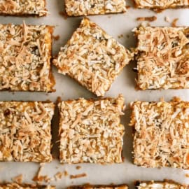 A batch of golden-brown, coconut-topped bars arranged on a white surface, with one square slightly separated from the others.