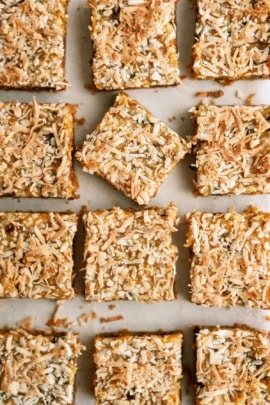 A batch of golden-brown, coconut-topped bars arranged on a white surface, with one square slightly separated from the others.