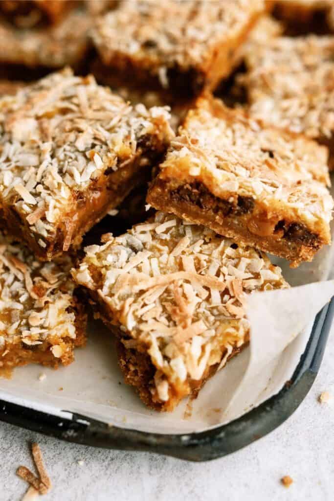 Close-up of a tray with several layered dessert bars topped with shredded coconut, revealing a caramel and chocolate filling. The tray is lined with parchment paper.