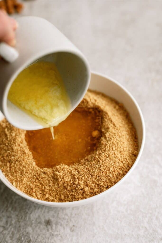 A hand pouring melted butter from a white cup into a bowl of crushed graham crackers.