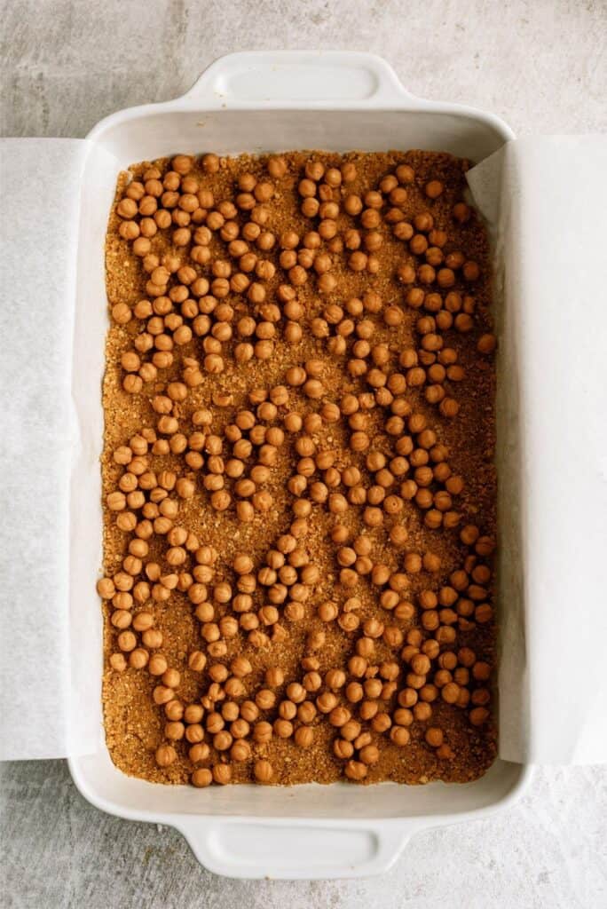 A baking dish lined with parchment paper, filled with an even layer of pastry dough and topped with numerous brown baking beans, ready for blind baking.