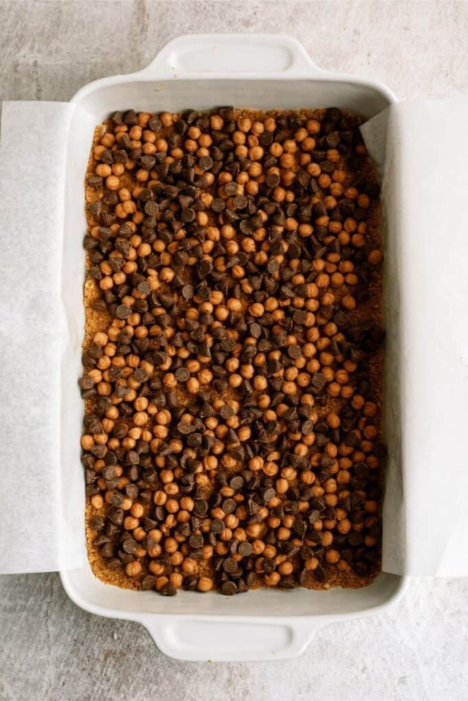 A baking dish filled with an even layer of chocolate and peanut butter chips on a crust, lined with parchment paper.