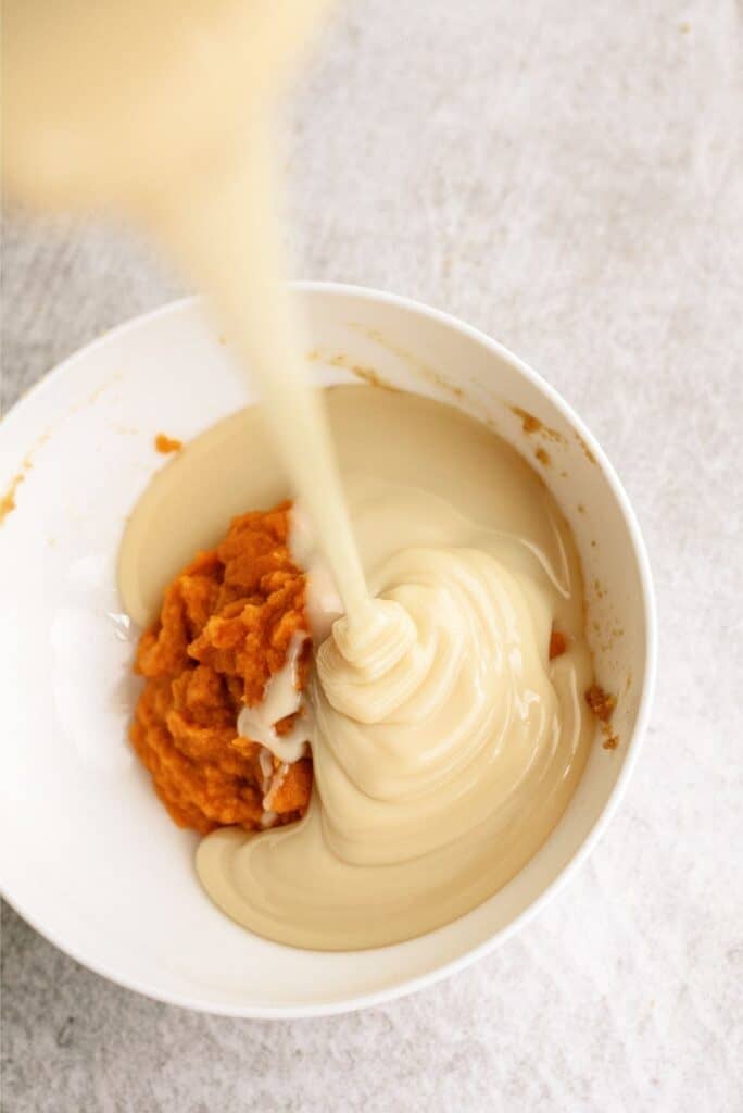 A white bowl contains mashed orange pumpkin and is having a thick, light-colored sauce being poured into it. The background is a light, textured surface.