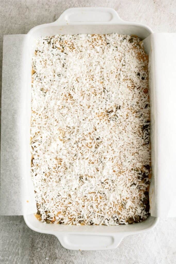 A rectangular white baking dish filled with a dessert topped with a layer of shredded coconut. The dish is lined with parchment paper.